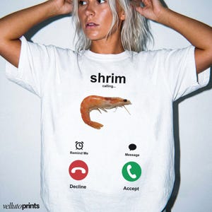 May include: White t-shirt featuring a cooked shrimp graphic with the text "shrimp calling...". Below the shrimp are phone icons for "Remind Me", "Decline", "Message", and "Accept". The brand name "vellutoprints" is at the bottom.