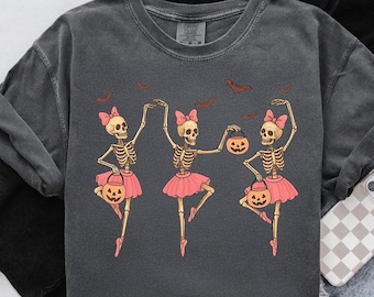Può includere: Felpa grigio scuro con un design a tema Halloween. Presenta tre ballerine scheletro con tutù e fiocchi rosa, che tengono lanterne a forma di zucca. Pipistrelli marroni volano sopra. La felpa ha un aspetto vintage e lavato.