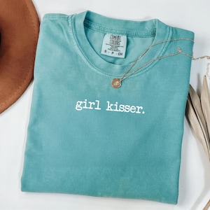 Puede incluir: Una camiseta Comfort Colors de color verde azulado con el texto "girl kisser" en blanco. Un collar dorado con un colgante está drapeado sobre la camiseta. Un sombrero marrón y una planta seca están en el fondo.