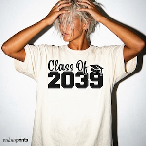 May include: A cream-coloured t-shirt displaying the text "Class Of 2039" in black, with a graduation cap graphic. The shirt has short sleeves and a crew neck. The brand name "vellutoprints" is at the bottom left.