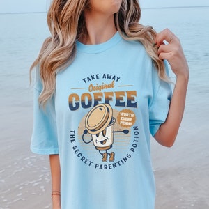 Op de afbeelding: Een lichtblauw t-shirt met een afbeelding van een koffiekopje met een gezicht en de tekst "Take Away Original Coffee The Secret Parenting Potion Worth Every Penny".