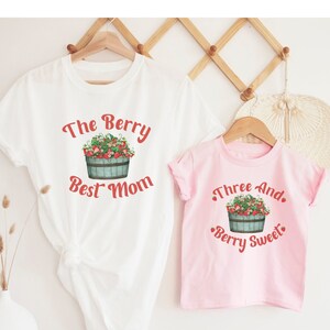May include: Two white t-shirts with a strawberry design. The larger shirt says "The Berry Best Mom" and the smaller shirt says "Three And Berry Sweet".