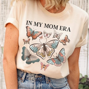 K&ouml;nnte beinhalten: Ein beiges T-Shirt mit dem Aufdruck "IN MY MOM ERA" in Schwarz. Das Shirt zeigt ein farbenfrohes Schmetterlingsdesign mit mehreren Schmetterlingen in verschiedenen Blaut&ouml;nen, Rosa und Orange.