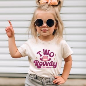 May include: A toddler girl wearing a white t-shirt with a pink and purple graphic that says "Two Rowdy Too Cute" and a pink cowboy hat. She is wearing denim shorts and sunglasses.