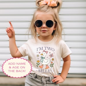 May include: A natural-colored toddler t-shirt with the text "I'M TWO SILLY!" and a cartoon goose design. The design includes flowers, a bow, and the option to add a name and age on the back. The word "NATURAL" is printed at the bottom.