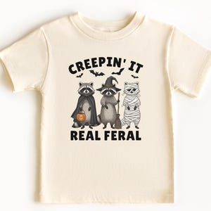 Op de afbeelding: Een crèmekleurig T-shirt met een Halloween-thema. Het ontwerp toont drie wasberen verkleed als heks, mummie en vampier, met de tekst "CREEPIN' IT REAL FERAL". Het shirt is gemaakt van zacht materiaal.