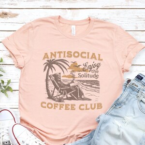 May include: A pink t-shirt featuring the text "ANTISOCIAL Enjoy the Solitude COFFEE CLUB" and a graphic of a skeleton sitting on a beach chair with a cup of coffee. The skeleton is wearing a hooded robe and has a skull face. The graphic is in brown and black.