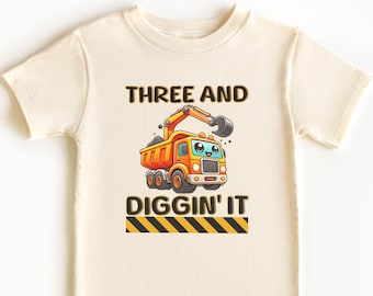 May include: Cream-colored t-shirt featuring a cartoon dump truck with the text "THREE AND DIGGIN' IT". The truck is orange and yellow with a smiling face. Below the text is a black and yellow striped design.