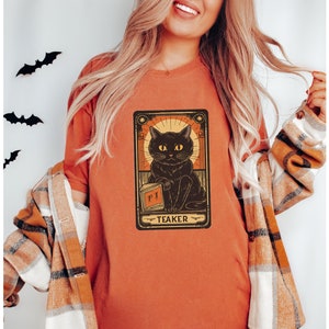 May include: A burnt orange t-shirt with a black cat illustration. The cat is holding a book with the word "PI" on it. The illustration is surrounded by a gold frame with the word "TEAKER" at the bottom.