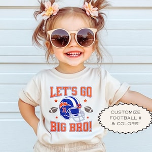 Puede incluir: Una camiseta beige para ni&ntilde;os peque&ntilde;os con el texto "LET'S GO BIG BRO!" en naranja y azul, con un casco de f&uacute;tbol americano y balones de f&uacute;tbol. La camiseta se combina con pantalones cortos beige. La imagen tambi&eacute;n incluye el texto "PERSONALIZA EL F&Uacute;TBOL # & COLORES!"