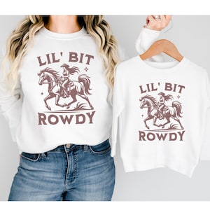 May include: White sweatshirts with a brown graphic of a cowgirl riding a horse, the text "LIL' BIT ROWDY" is above and below the graphic. One sweatshirt is worn, the other is on a wooden hanger.