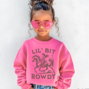 May include: A pink sweatshirt with the text "LIL' BIT ROWDY" and a graphic of a cowgirl riding a horse. The sweatshirt is a vibrant pink color and is paired with blue jeans. The words "SAFETY PINK" are printed at the bottom.