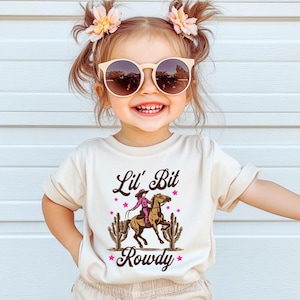 May include: A toddler wearing a beige t-shirt with a graphic of a girl riding a horse and the text "Lil' Bit Rowdy". The toddler is wearing beige shorts and has brown hair with pink flowers in it. The toddler is wearing sunglasses.