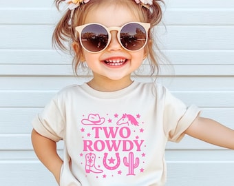 May include: A young child wearing a beige t-shirt and shorts, with sunglasses and floral hair accessories. The t-shirt features the text "TWO ROWDY" in pink, along with cowboy-themed graphics. The child is smiling and standing in front of a white background.