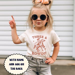 May include: A young child wearing a cream-colored t-shirt with the words "THREE & ROWDY" and a graphic of a cowboy and cowgirl riding a horse. The shirt also has the text "WITH NAME AND AGE ON THE BACK". The child is wearing sunglasses and denim shorts.