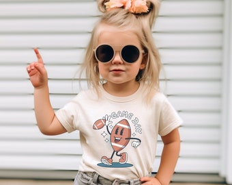 May include: A young child wearing a beige t-shirt with a cartoon football graphic and the text "Game Day". The child is also wearing denim shorts and sunglasses. The background is a white slatted wall.