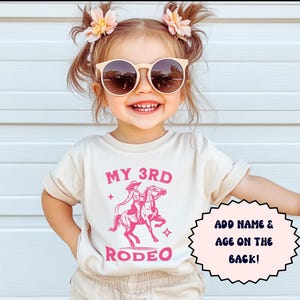 May include: A young child wearing a light beige t-shirt with the text "My 3rd Rodeo" and a pink graphic of a cowgirl riding a horse. The shirt has a pink star on each side of the text. The child is wearing a pair of brown shorts and sunglasses. The text "Add name & age on the back!" is in a pink circle on the right side of the image. The text "NATURAL" is at the bottom of the image.