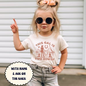 Puede incluir: Una camiseta beige claro con un gr&aacute;fico marr&oacute;n de una vaquera con un lazo y el texto "This gal is two rowdy". La camiseta tiene mangas cortas y cuello redondo. El texto "With name & age on the back" est&aacute; en un c&iacute;rculo blanco en el lado izquierdo de la imagen. El texto "NATURAL" est&aacute; en el lado derecho de la imagen.