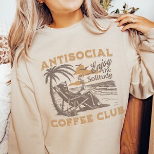 May include: A beige sweatshirt with a graphic print of a skeleton sitting on a beach chair with a palm tree and the text "ANTISOCIAL Enjoy the Solitude COFFEE CLUB".
