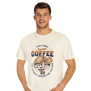 Op de afbeelding: Een wit T-shirt met een bruine en blauwe afbeelding van een koffiebeker met de tekst "Take Away Original Coffee The Secret Parenting Potion Worth Every Penny".