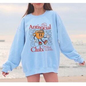 K&ouml;nnte beinhalten: Hellblauer Pullover mit einem Grafikdruck einer Comic-Kaffeetasse mit dem Text "The Antisocial Coffee Club. Espresso yourself, silently."