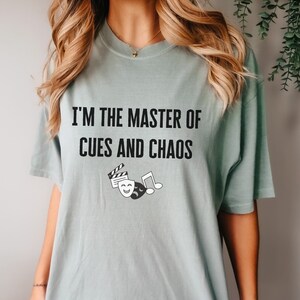 May include: A light blue t-shirt with the text "I'm the master of cues and chaos" in black. The shirt features a graphic of a film clapboard, a musical note, and a theater mask.