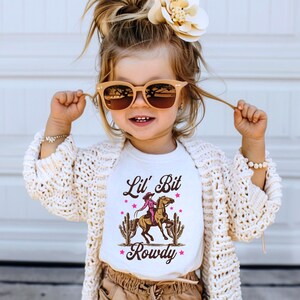 K&ouml;nnte beinhalten: Ein wei&szlig;es T-Shirt mit einem Aufdruck einer reitenden M&auml;dchen mit dem Text "Lil' Bit Rowdy" in braunen und rosa Buchstaben.