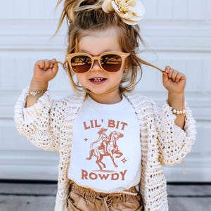 May include: A young girl wearing a white t-shirt with a brown graphic of a cowgirl riding a horse and the text "Lil' Bit Rowdy". She is wearing a white cardigan, brown shorts, and sunglasses.