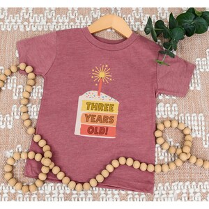 May include: A maroon toddler t-shirt with a graphic of a layered cake with a lit sparkler on top. The cake has the text "THREE YEARS OLD!" on it.