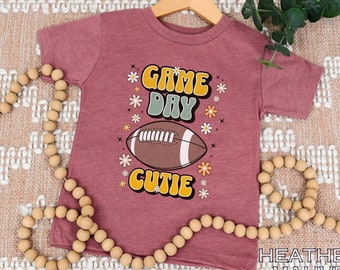 May include: A heather mauve t-shirt with a retro-style graphic of a football and the text "Game Day Cutie".