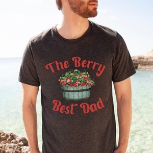 May include: A dark grey t-shirt with the text "The Berry Best Dad" in red lettering. A cartoon illustration of a bucket overflowing with red strawberries is above the text.