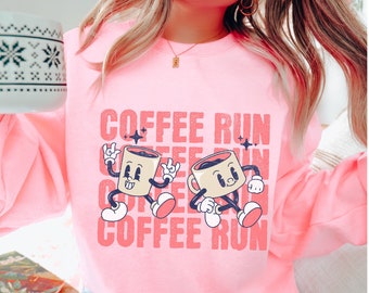 Op de afbeelding: Roze sweatshirt met een afbeelding van twee cartoon koffiekopjes die rennen en de tekst "COFFEE RUN" die wordt herhaald.