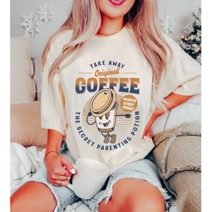 Op de afbeelding: Een lichtgeel T-shirt met een retro-grafische afbeelding van een koffiekopje met de tekst "Take Away Original Coffee The Secret Parenting Potion Worth Every Penny".