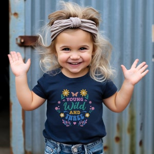 May include: A young girl wearing a navy blue t-shirt with the text "Young Wild and Three" in colorful letters surrounded by flowers and butterflies. She is smiling and has blonde hair with a light brown headband.