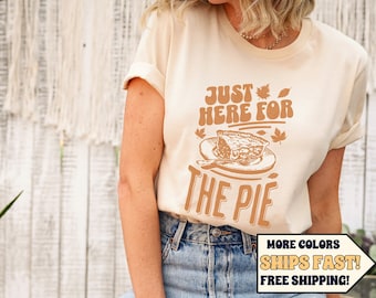 May include: Cream-colored t-shirt with the text "JUST HERE FOR THE PIE" in brown. The graphic includes a pie slice on a plate with fall leaves. The shirt is paired with blue jeans.