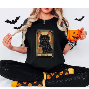 May include: A black t-shirt with a vintage tarot card design featuring a black cat holding a potion bottle. The card reads "Teaker" and is surrounded by a gold frame.