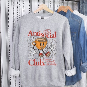 K&ouml;nnte beinhalten: Grauer Pullover mit einem Comic-Kaffeebecher-Motiv und dem Text "The Antisocial Club. Espresso yourself, silently."