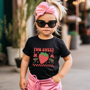 May include: A black t-shirt with a red strawberry graphic and the text "TWO SWEET" in white. The shirt is worn by a child with pink hair and sunglasses.