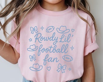 May include: A light pink t-shirt with the text "Rowdy Lil' football fan" in light blue. The design includes cowboy hats, footballs, bows, stars, and hearts. The shirt is made of soft material.