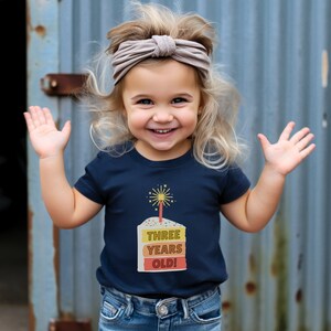 May include: A young child wearing a navy blue t-shirt with a birthday cake graphic and the text "THREE YEARS OLD!" The child is smiling and has blonde hair with a light brown headband.