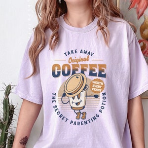 Op de afbeelding: Paars T-shirt met een retro-grafische afbeelding van een koffiebeker met de tekst "Take Away Original Coffee The Secret Parenting Potion Worth Every Penny".