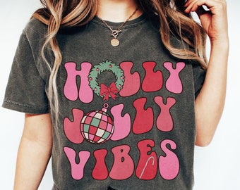 May include: A dark gray t-shirt with a pink and red retro design that says "HOLLY JOLLY VIBES". The design includes a wreath with a red bow and a red and green ornament.