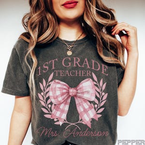 Puede incluir: Camiseta gris oscuro con el texto "1st Grade Teacher" en rosa, sobre un lazo rosa a cuadros y un diseño de corona de laurel. El nombre "Mrs. Anderson" está impreso debajo. La camiseta es de estilo casual.