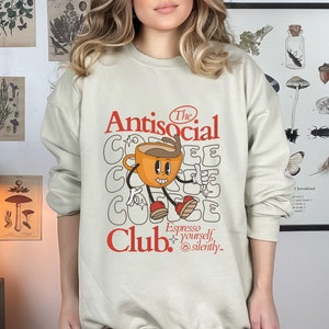 K&ouml;nnte beinhalten: Ein beiger Pullover mit einem Grafikdesign, das eine Comic-Kaffeetasse mit dem Text "The Antisocial Club. Espresso yourself, silently." zeigt.