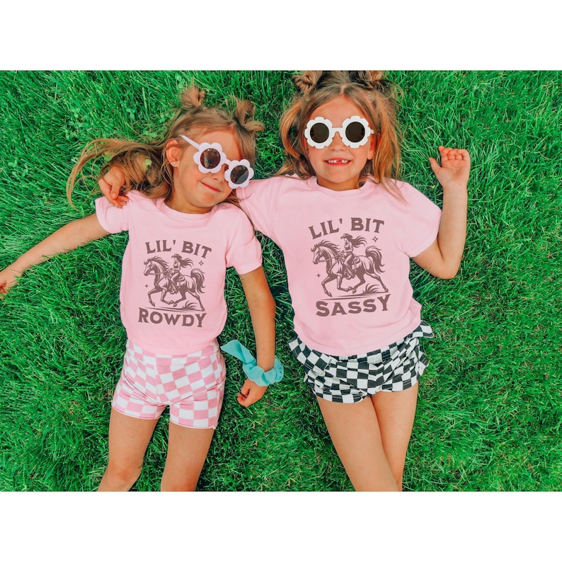 May include: Two young children wearing matching pink t-shirts with a horse and rider graphic and the text "LIL' BIT ROWDY" and "LIL' BIT SASSY". They are also wearing sunglasses and checkered shorts, lying on green grass.