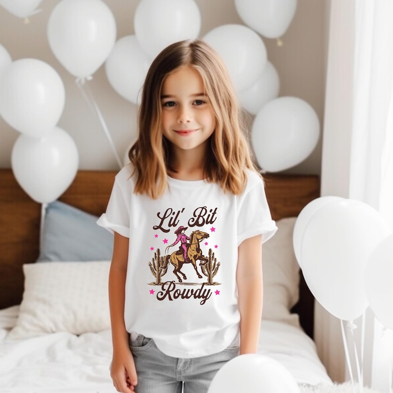 May include: A white t-shirt with a pink and brown graphic of a girl riding a horse with the text "Lil' Bit Rowdy" printed on it.