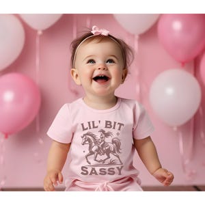 May include: A light pink t-shirt with the text "LIL' BIT SASSY" and a graphic of a cowgirl riding a horse. The baby is smiling and wearing a pink bow headband. Pink and white balloons are in the background.