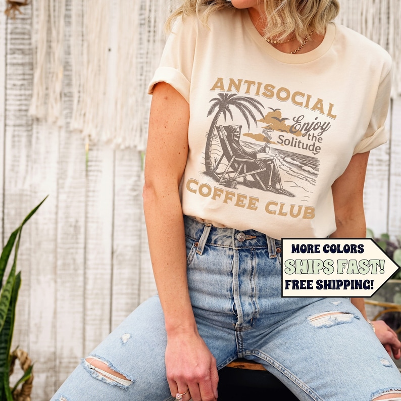 May include: A beige t-shirt featuring a graphic of a skeleton relaxing on a beach chair beneath a palm tree. The text on the shirt reads "Antisocial Enjoy the Solitude Coffee Club".