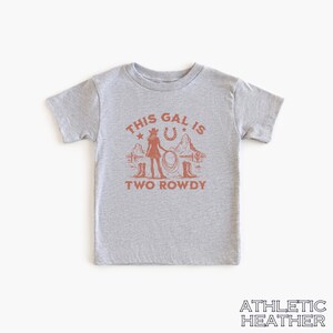 Puede incluir: Camiseta gris de manga corta con un gr&aacute;fico de una vaquera sosteniendo un lazo frente a una herradura y estrellas. El texto "This Gal Is Two Rowdy" est&aacute; impreso debajo del gr&aacute;fico.