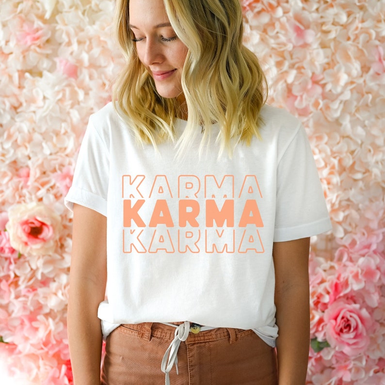 Może przedstawiać: Biały t-shirt z napisem "KARMA" powt&oacute;rzonym trzy razy w kolorze brzoskwiniowo-pomarańczowym. Tekst jest ułożony pionowo i obwiedziony białym kolorem.